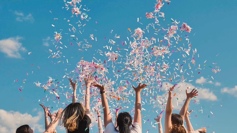 Friends,Celebrating,Joyfully,Outdoors,,Throwing,Colorful,Confetti,Under,A,Blue